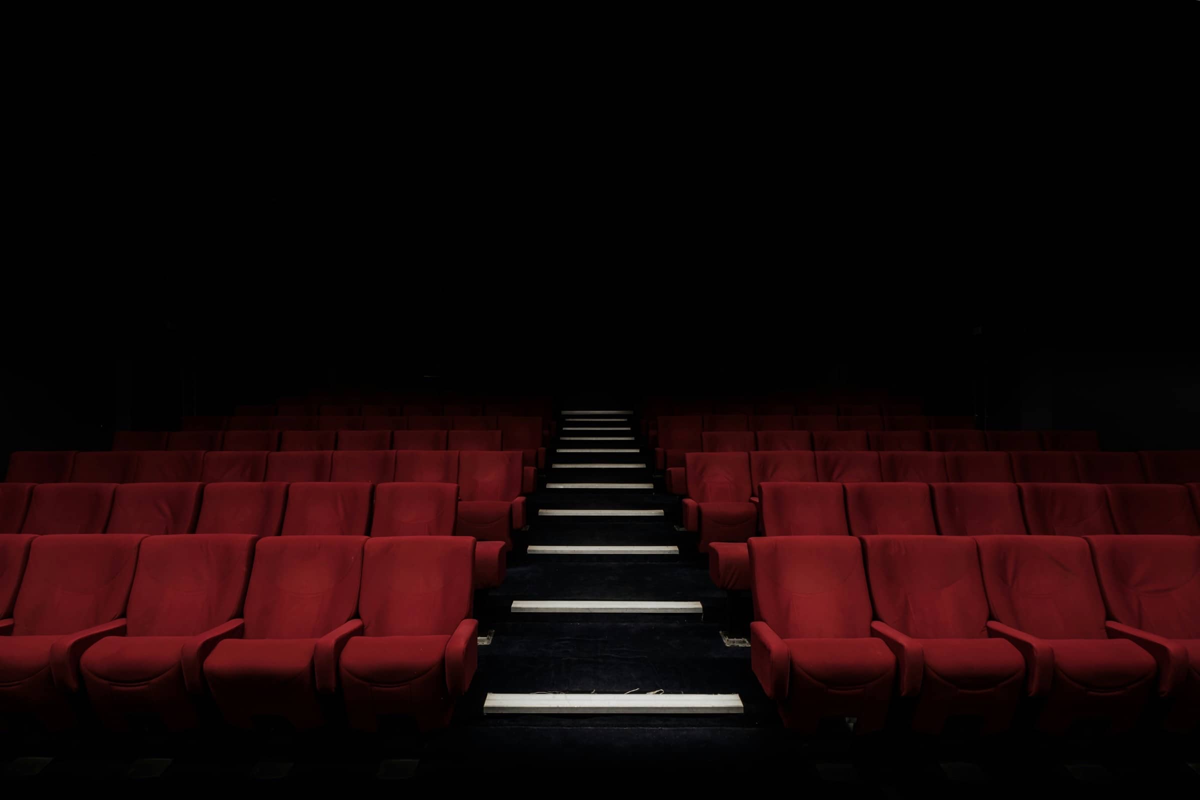 Sala de cine vacía con butacas rojas y una escalera central iluminada que sube hacia la salida.