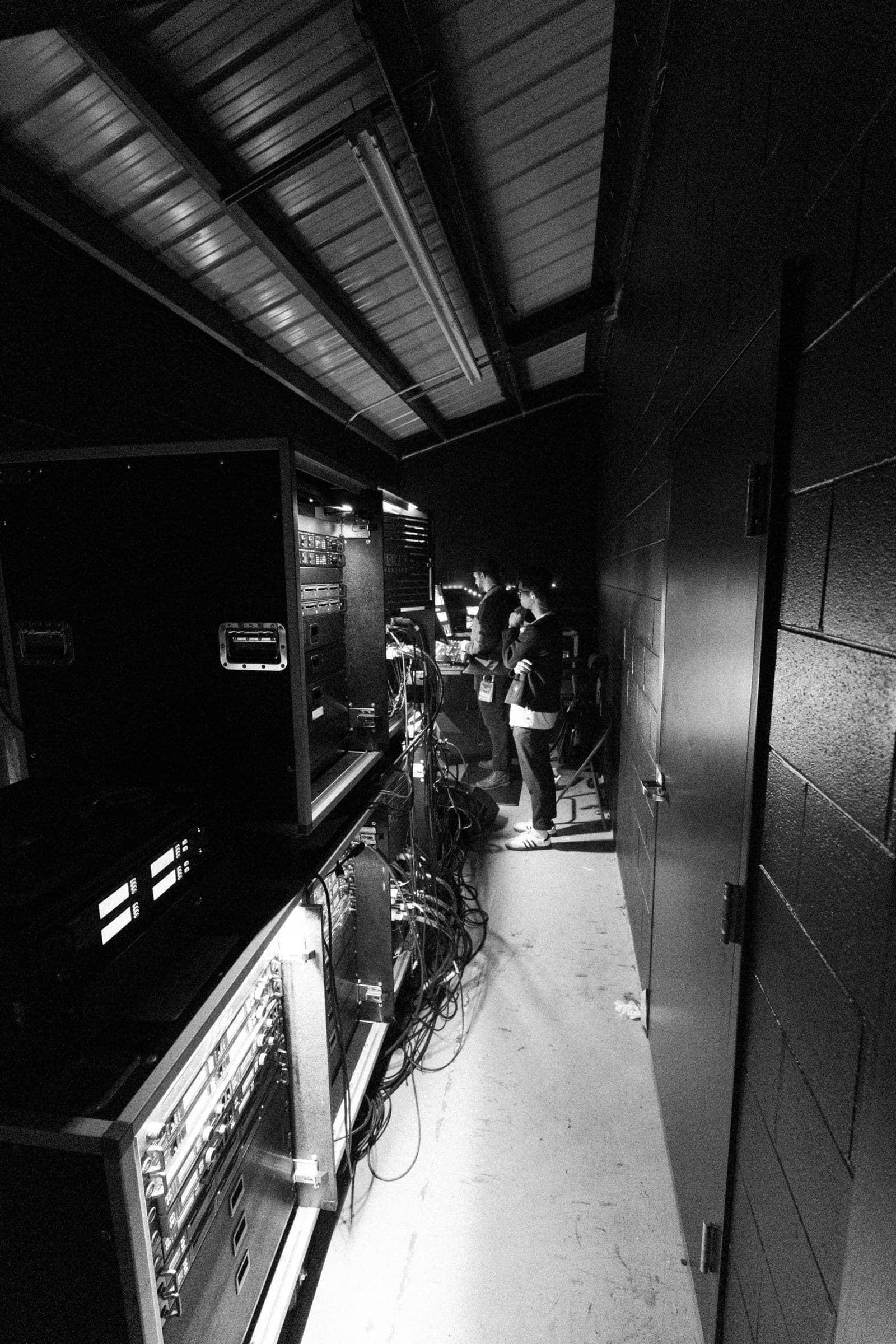 Pasillo técnico con un rack de material de sonido y personal trabajando al fondo, en blanco y negro.