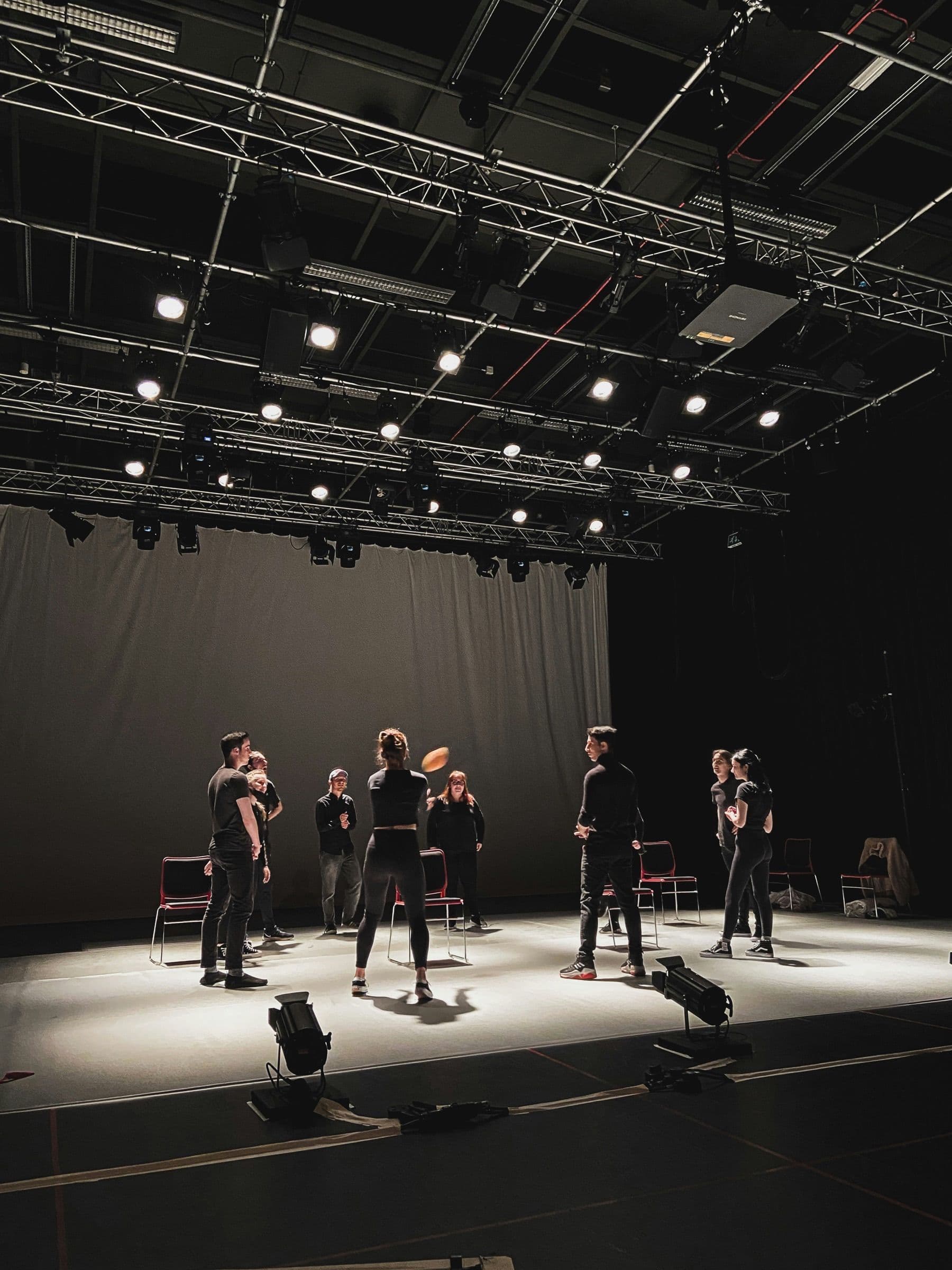Assaig de teatre en un escenari amb estructures de llums i intèrprets asseguts en cercle.